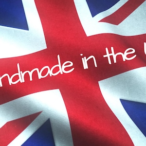May include: A fabric Union Jack flag featuring the red, white, and blue colors of the United Kingdom. The text "Handmade in the UK" is printed in a white, rounded, sans-serif font across the center of the flag, which has a wavy texture.