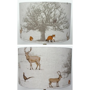 May include: A cylindrical lampshade featuring a light grey fabric base with a printed woodland scene. The design includes a large central tree, a brown fox, a squirrel, an owl, and several deer. A pheasant is also visible on the lower section.