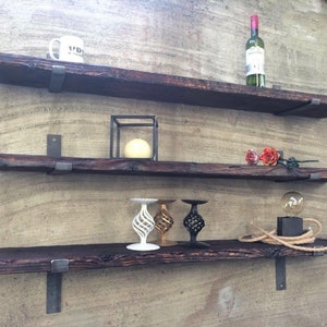 May include: Three rustic, dark wood floating shelves mounted on a textured wall. The shelves are supported by metal brackets. Various decorative items are displayed, including a wine bottle, a mug, candles, and decorative candle holders.