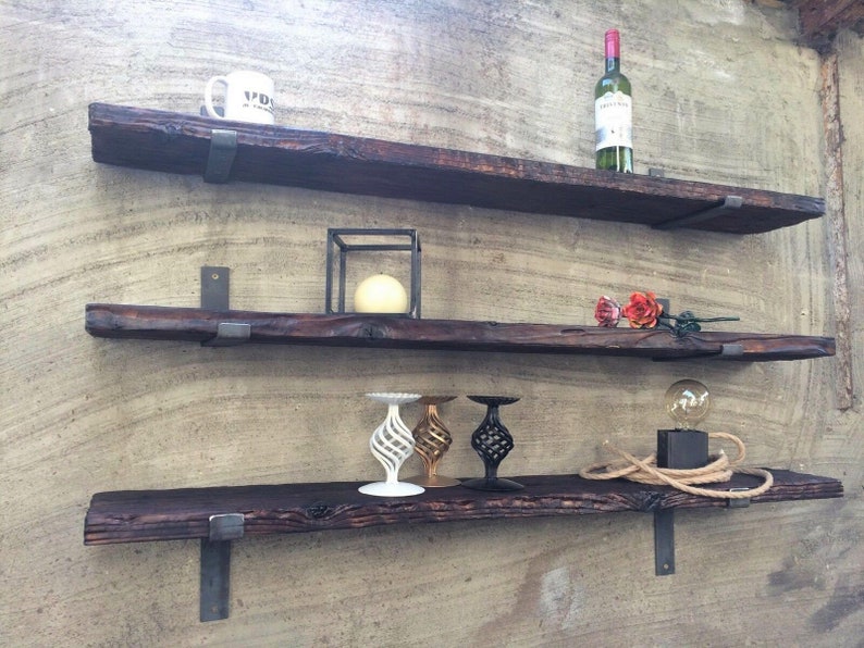 May include: Three rustic, dark wood floating shelves mounted on a textured wall. The shelves are supported by metal brackets. Various decorative items are displayed, including a wine bottle, a mug, candles, and decorative candle holders.