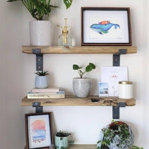 May include: Three-tiered wooden shelves with various decorative items. The top shelf holds a potted plant, a framed watercolor, and a glass spray bottle. The middle shelf displays a small succulent, books, and a potted plant. The bottom shelf features a framed watercolor and a decorative plant.