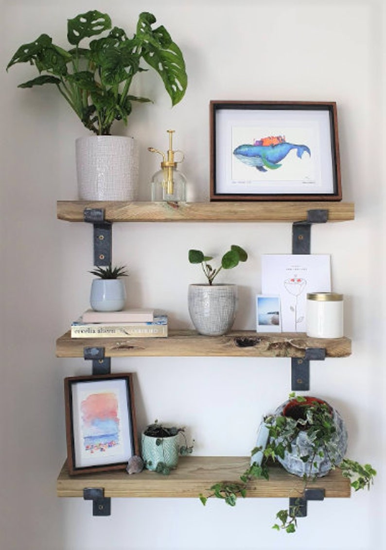 May include: Three-tiered wooden shelves with various decorative items. The top shelf holds a potted plant, a framed watercolor, and a glass spray bottle. The middle shelf displays a small succulent, books, and a potted plant. The bottom shelf features a framed watercolor and a decorative plant.