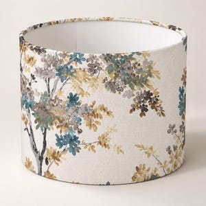 May include: A cylindrical drum lampshade featuring a cream-colored fabric base with a watercolor-style botanical print. The pattern displays tree branches with clusters of leaves and small flowers in shades of teal, muted yellow, and gray.