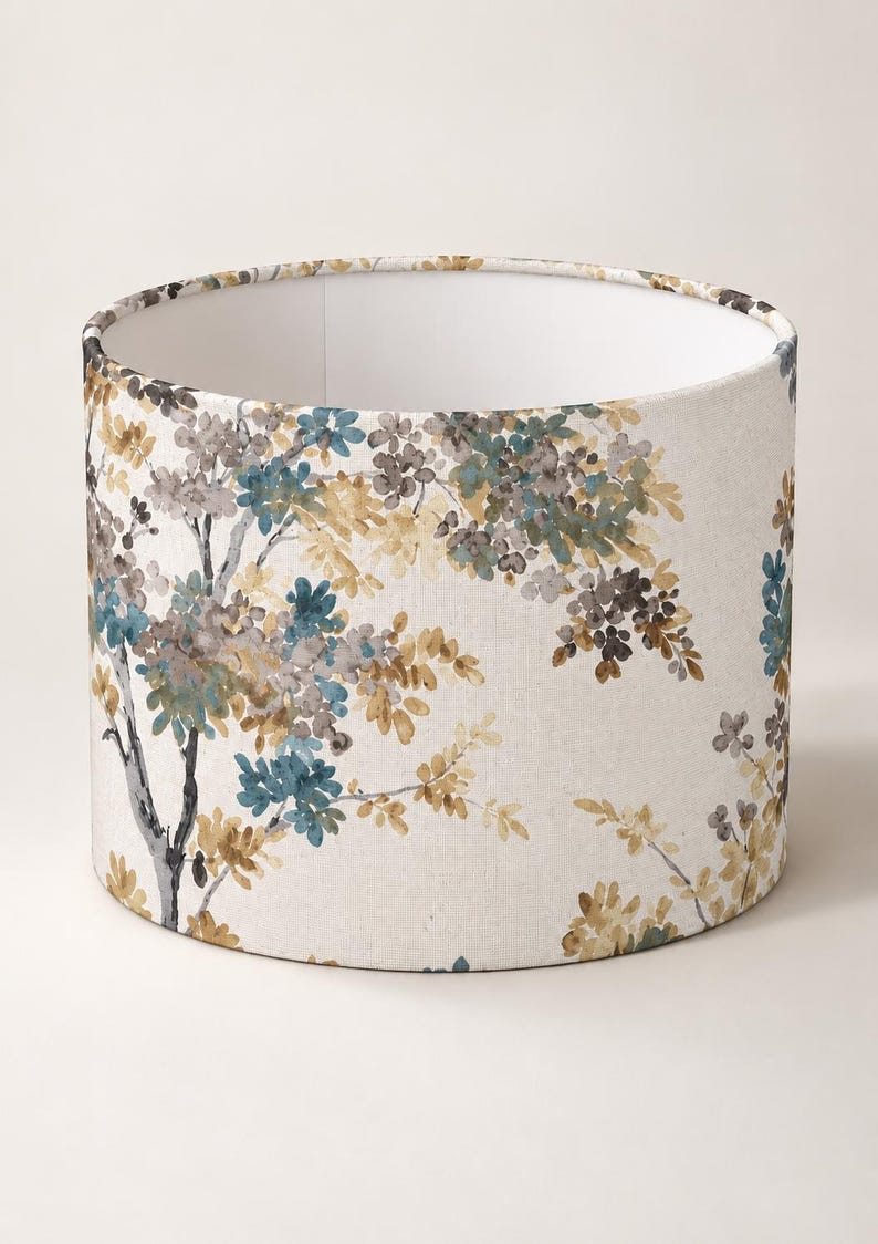May include: A cylindrical drum lampshade featuring a cream-colored fabric base with a watercolor-style botanical print. The pattern displays tree branches with clusters of leaves and small flowers in shades of teal, muted yellow, and gray.