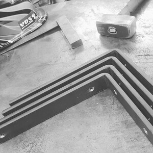 May include: Black and white image of metal L-shaped brackets with pre-drilled holes, stacked on a metal surface. A hammer, a metal square, and a welding mask are also visible in the background, suggesting a metalworking theme.
