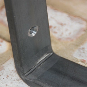 May include: Close-up of a dark gray metal bracket with a right-angle bend. A single hole is drilled near the top edge. The bracket appears to be made of heavy-duty steel, suitable for furniture or shelving.