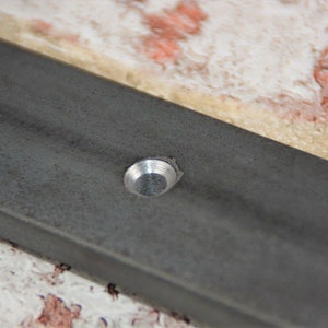May include: Close-up of a section of a dark gray metal frame with a silver rivet. The frame has a slightly textured surface and is set against a background of beige and red-toned tiles.