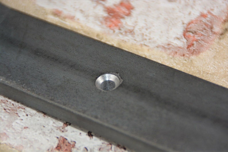 May include: Close-up of a section of a dark gray metal frame with a silver rivet. The frame has a slightly textured surface and is set against a background of beige and red-toned tiles.