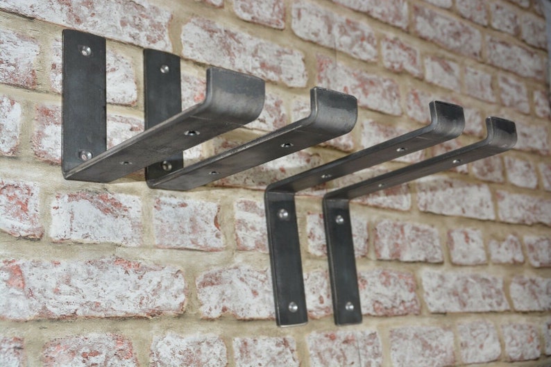 May include: Set of three industrial-style metal shelf brackets. The brackets are a dark gray color and are mounted on a brick wall. Each bracket has a right-angle design with pre-drilled holes for mounting. The brackets are designed to support shelves.