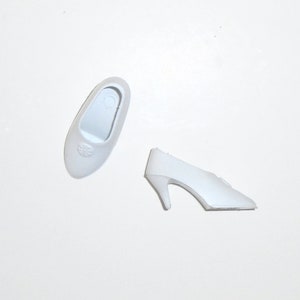 May include: Two pairs of white plastic doll shoes. One pair is a flat, closed-toe shoe. The other pair is a high-heeled pump.