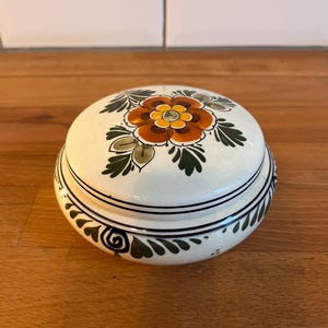 May include: A round ceramic trinket box with a lid. The white box features a floral design on the lid, with a brown and yellow flower and green leaves. The rim has a black and green decorative pattern.