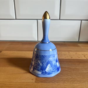 May include: A blue and white ceramic bell with a gold-coloured top. The bell features a detailed landscape scene in blue. The handle is light blue, and the bell is sitting on a wooden surface.