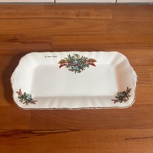 May include: A rectangular white ceramic serving tray with gold trim and decorative floral and plaid accents. The tray has the text "St. Mary's Loch" printed on it. The tray is on a wooden surface.