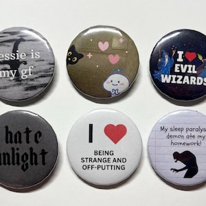 May include: Six circular badges with different designs and text. The badges display phrases such as "nessie is my gf," "I love evil wizards," and "I hate sunlight." Other badges feature illustrations of a cat, a cloud, and a monster.