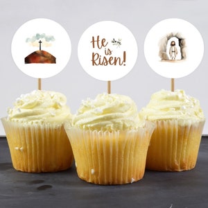 May include: Three cupcakes with white frosting and sprinkles topped with round paper cupcake toppers. The toppers feature watercolour illustrations of a cross on a hill, the words "He is Risen!" and a figure walking out of a cave.
