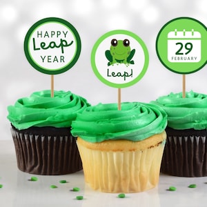 Puede incluir: Tres cupcakes verdes con glaseado verde cubiertos con adornos redondos de papel para cupcakes. Los adornos representan una rana, un calendario con la fecha 29 de febrero y el texto "Happy Leap Year".