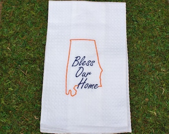 May include: A white waffle-weave kitchen towel featuring an orange embroidered outline of the state of Alabama. Centered within the state outline is the dark blue embroidered text Bless Our Home in a cursive script font.