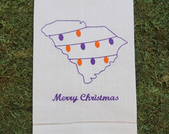 May include: A white rectangular hand towel featuring an embroidered outline of the state of South Carolina in purple thread. The state shape is decorated with purple and orange Christmas light bulbs. The text Merry Christmas is embroidered below.