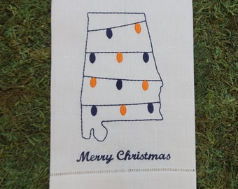 May include: A white rectangular tea towel featuring an embroidered outline of the state of Alabama decorated with navy blue and orange Christmas light bulbs. The text Merry Christmas is embroidered in navy blue below the state outline.