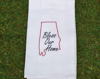 May include: A white waffle-weave kitchen towel featuring an embroidered red outline of the state of Alabama. Inside the state outline, the text Bless Our Home is stitched in black cursive lettering. The fabric has a textured, grid-like pattern.