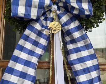 May include: A large decorative bow featuring a blue and white gingham check pattern. The bow is accented with a gold-colored football helmet charm hanging from a thin gold chain and a plain white ribbon streamer centered below the knot.