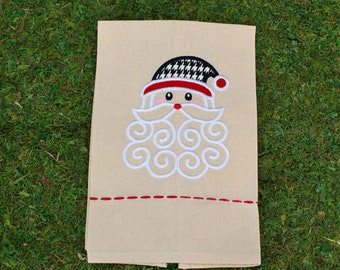 May include: A beige rectangular kitchen towel featuring an embroidered Santa Claus face. The Santa has a black and white houndstooth hat with a red brim, a white swirled beard, and a red nose. A horizontal red dashed line sits below the face.