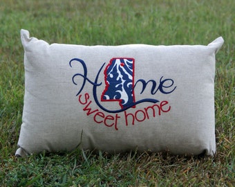 May include: A beige pillow with the words "Home sweet home" embroidered in red and blue thread. The state of Alabama is embroidered in blue and white in the center of the pillow.