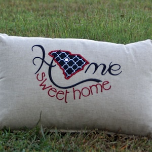 May include: A rectangular beige pillow embroidered with the words "Home sweet home." The word "Home" is in a cursive font, with the "O" replaced by a map of South Carolina in red, black, and white.
