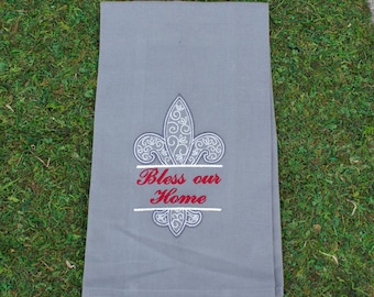 May include: A rectangular gray kitchen towel featuring an embroidered white fleur-de-lis design with intricate scrollwork patterns. Below the fleur-de-lis, the text "Bless our Home" is stitched in red thread, centered between two horizontal lines.