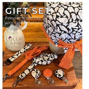 May include: A gift set featuring a ponytail scrub cap, badge reel, lanyard, and keychain. The items have a black and white ghost pattern with orange polka dot accents. The text "GIFT SET" and "LOLLI CAPS" are visible.