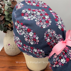 May include: Navy blue ponytail scrub cap patterned with red, pink, and white paw prints. The cap has a pink bow and is designed to be worn during medical procedures. The text "Women's Ponytail Scrub Cap" is visible.