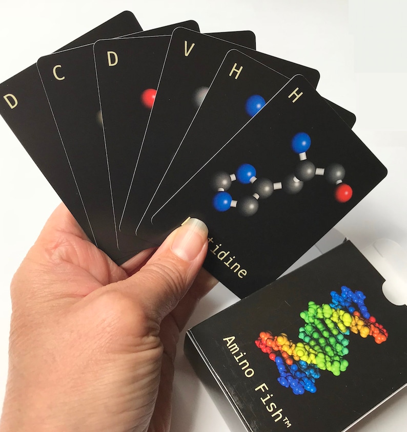 May include: A set of black cards with white letters and a molecular structure on one card. The cards are fanned out in a hand. The box for the cards is black with the text "Amino Fish&trade;" in white letters and a colorful DNA molecule graphic.