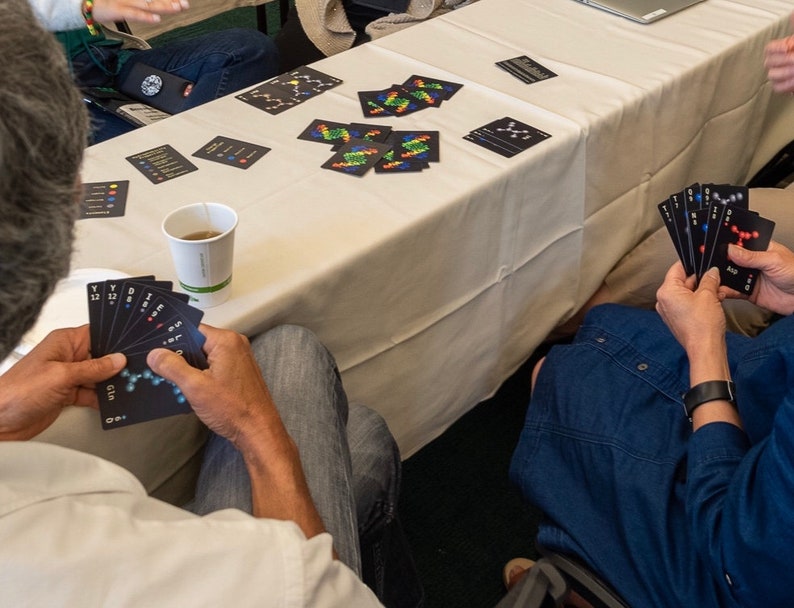 May include: People playing a card game with black cards featuring colorful molecular structures and numbers. The cards are laid out on a white tablecloth with a cup of tea on the table.