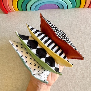 May include: A hand holds four small fabric pillows with different patterns: black and white diamonds, black and white stripes, white with black crosses, and a brown pillow with white polka dots. A wooden rainbow arch is in the background.
