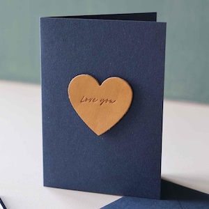 May include: A navy blue card with a tan leather heart. The words "Love you" are inscribed on the heart. A matching envelope is visible. The card is a romantic gesture.