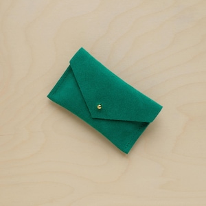 May include: A small, emerald green envelope-style wallet made of soft material. The wallet has a flap closure secured by a small, gold-colored stud. The wallet is displayed on a light wood surface.