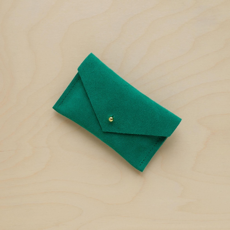 May include: A small, emerald green envelope-style wallet made of soft material. The wallet has a flap closure secured by a small, gold-colored stud. The wallet is displayed on a light wood surface.