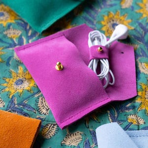 May include: A close-up of a fuchsia pink suede earbud case with a gold snap closure, holding white earbuds. Other colorful suede cases in teal, orange, and light blue are visible. The cases are on a teal floral patterned fabric.