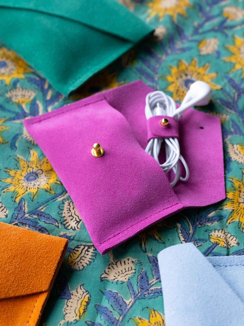 May include: A close-up of a fuchsia pink suede earbud case with a gold snap closure, holding white earbuds. Other colorful suede cases in teal, orange, and light blue are visible. The cases are on a teal floral patterned fabric.