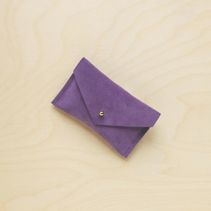 May include: A small, purple suede envelope-style wallet with a gold-colored snap closure. The wallet is rectangular with a flap closure and is set against a light wood grain background.
