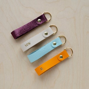 May include: Four leather keychains with gold rings and gold metal clasps. The keychains are in different colours: burgundy, white, light blue, and orange. The white keychain has the initials "MW" on it, the light blue keychain has no text, and the orange keychain has the letter "R" on it.
