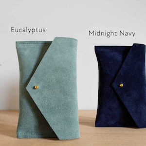 May include: Two suede pouches, one in eucalyptus green and the other in midnight navy blue, are displayed. Each pouch has a unique flap closure secured with a small gold-colored stud. The text "For Colour Reference: Eucalyptus, Midnight Navy" is visible.