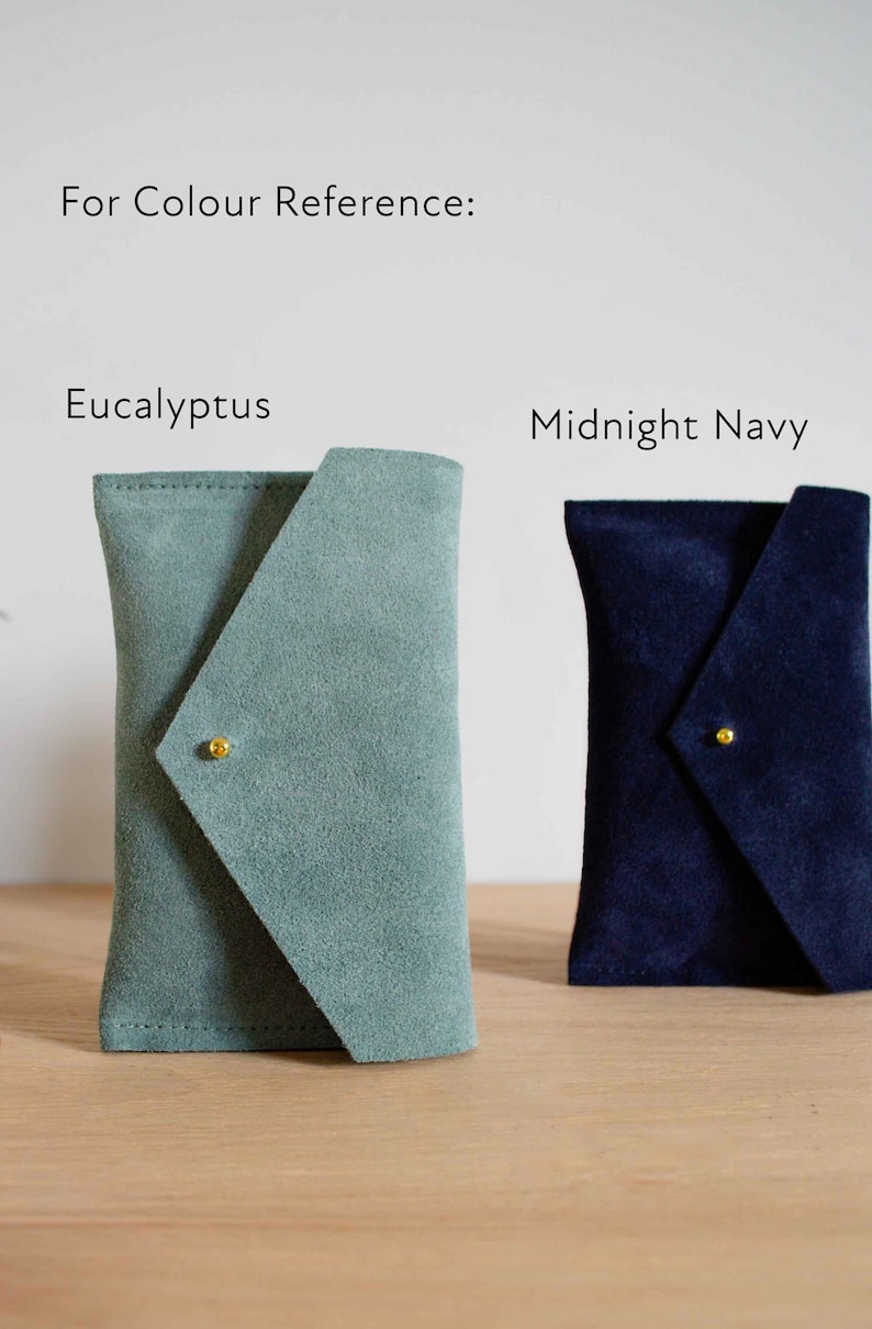 May include: Two suede pouches, one in eucalyptus green and the other in midnight navy blue, are displayed. Each pouch has a unique flap closure secured with a small gold-colored stud. The text "For Colour Reference: Eucalyptus, Midnight Navy" is visible.