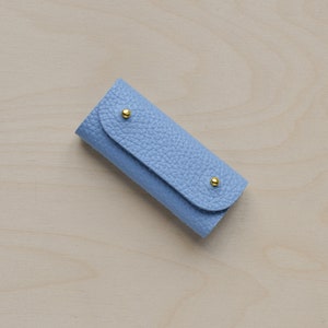 May include: A light blue leather key holder with gold accents. The key holder is closed and has a textured surface.