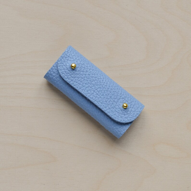 May include: A light blue leather key holder with gold accents. The key holder is closed and has a textured surface.