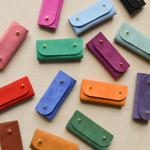 May include: Assortment of colourful suede keychains with gold accents. The keychains are arranged on a light wood surface.