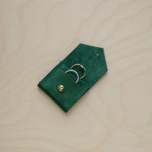 May include: A small green suede pouch with a flap and a button closure. The pouch is open and contains two rings, one gold and one silver.