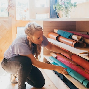 May include: A woman in a light purple striped shirt and dark pants is selecting from a shelf of rolled leather hides. The hides are various colors, including brown, teal, pink, and red. The wooden shelves are part of a larger wooden structure.
