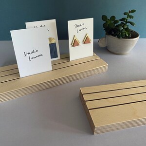 K&ouml;nnte beinhalten: Holz-Ausstellungsst&auml;nde mit Schlitzen, die wei&szlig;e Karten mit dem Text "Studio Lowen" halten. Ein St&auml;nder zeigt Ohrringe mit goldenen und rosa Dreiecksdesigns. Eine kleine Topfpflanze befindet sich im Hintergrund.