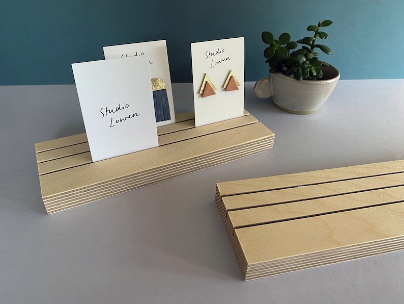 K&ouml;nnte beinhalten: Holz-Ausstellungsst&auml;nde mit Schlitzen, die wei&szlig;e Karten mit dem Text "Studio Lowen" halten. Ein St&auml;nder zeigt Ohrringe mit goldenen und rosa Dreiecksdesigns. Eine kleine Topfpflanze befindet sich im Hintergrund.